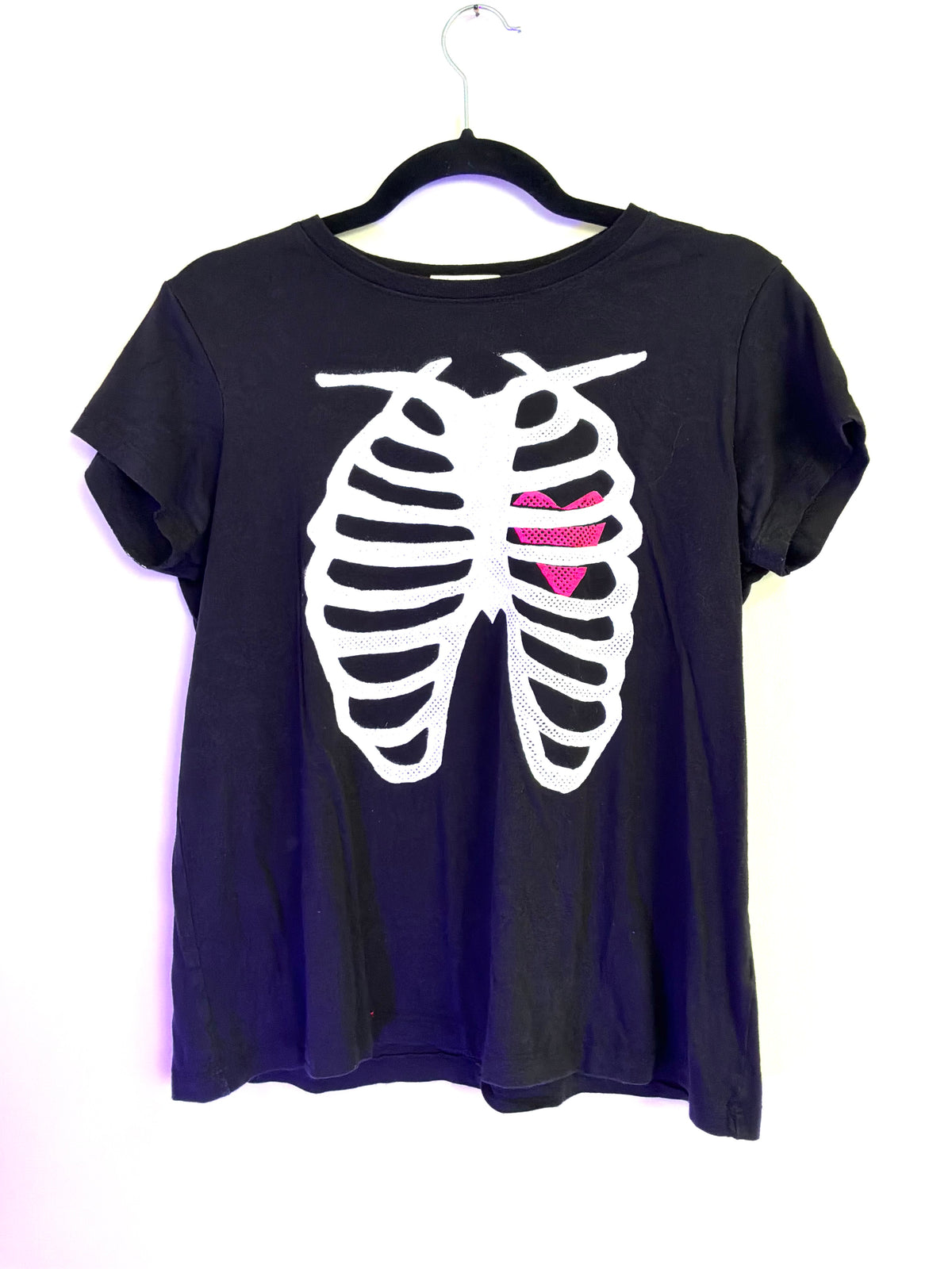 A black short-sleeve t-shirt hanging on a black hanger against a white background, featuring a white rib cage graphic with a pink heart shape inside the left rib cage.