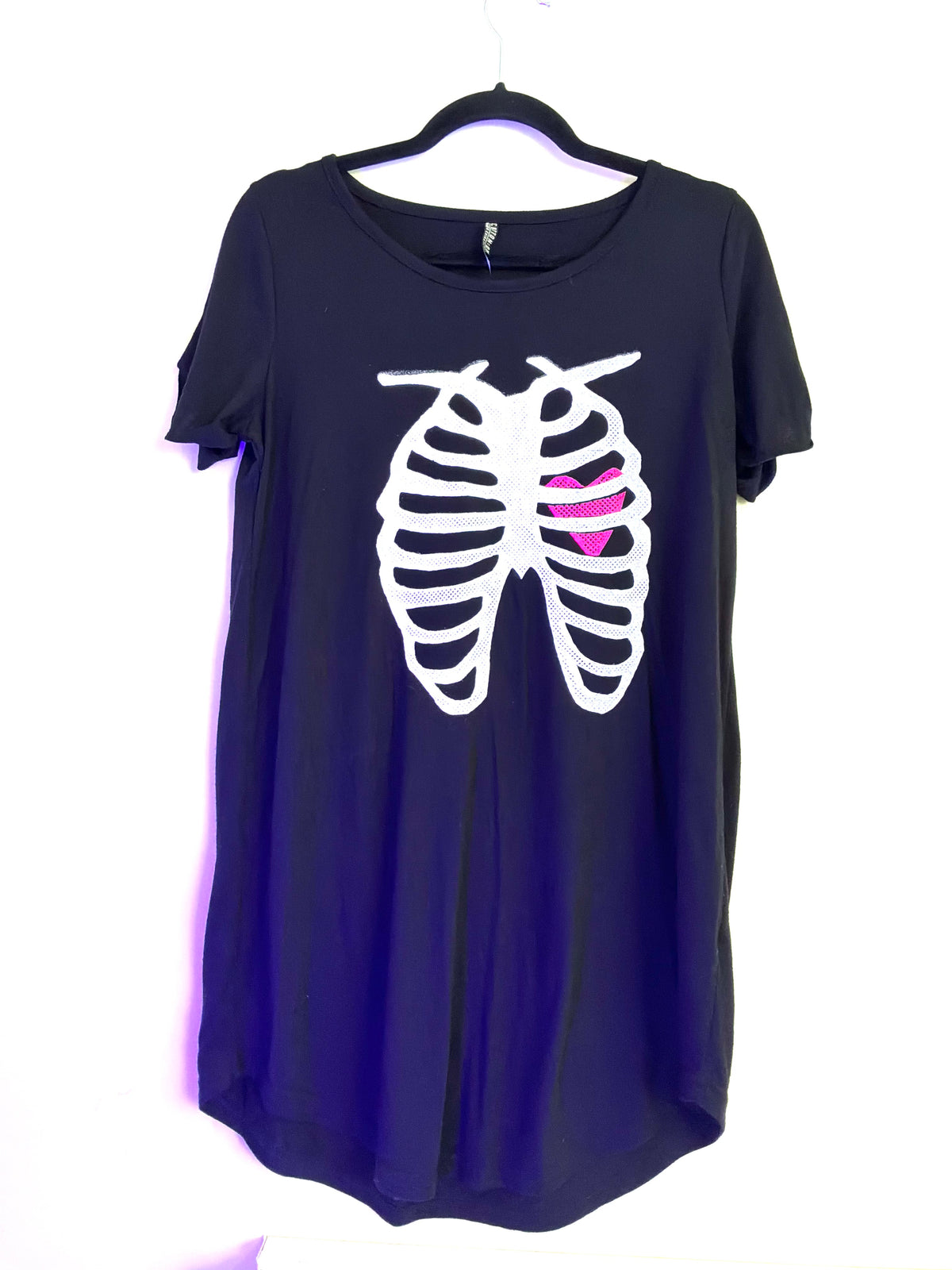 A black short-sleeve t-shirt hanging on a black hanger against a white background, featuring a white rib cage graphic with a bright pink heart in the center.